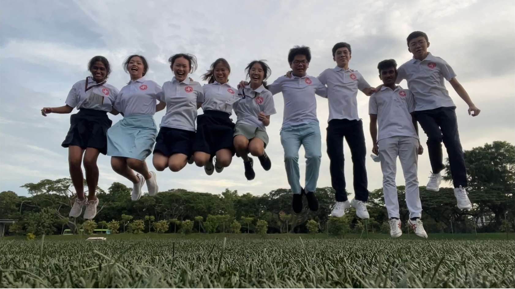 Eight students in white uniforms jump together laughing on a grass field with trees and cloudy sky behind them.